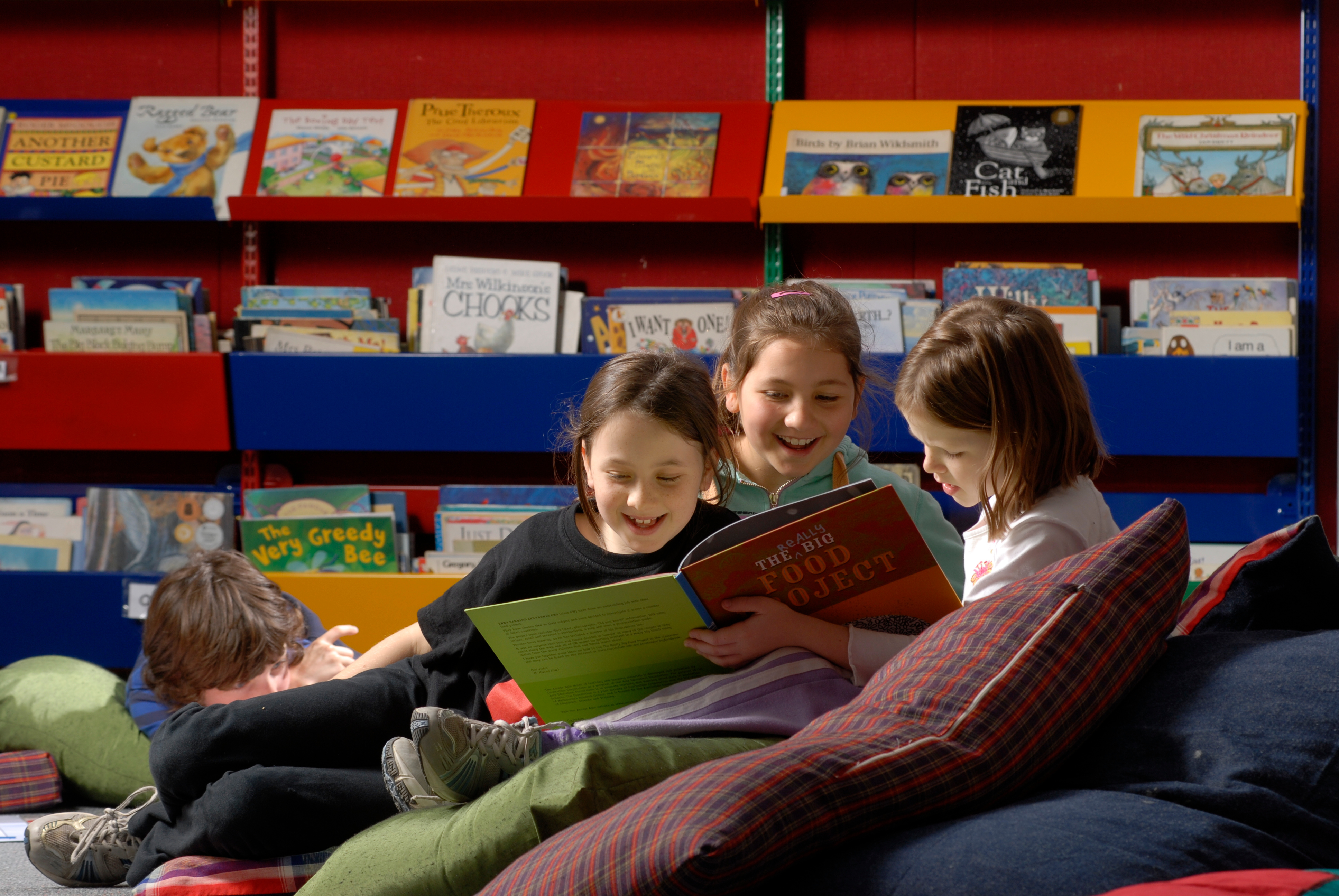 Three children in a library reading, sitting on a mound of pillows in from of book displays